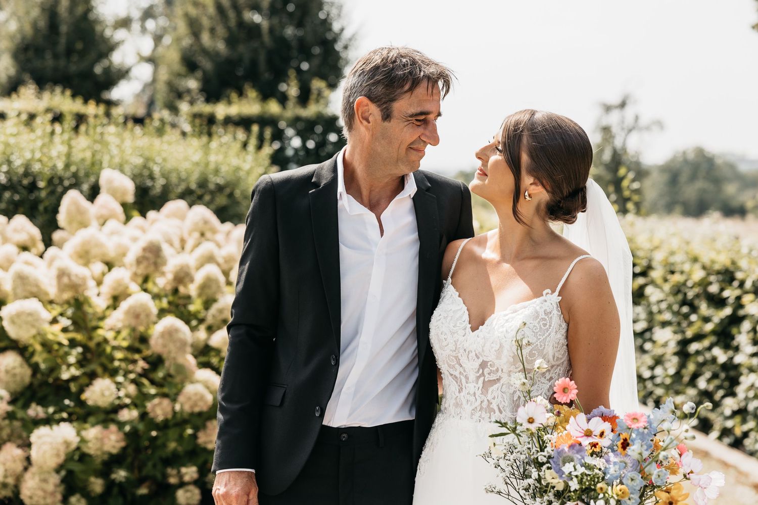 Bride and groom sharing tender glance while standing together near white hydrangeas in sunny garden setting.