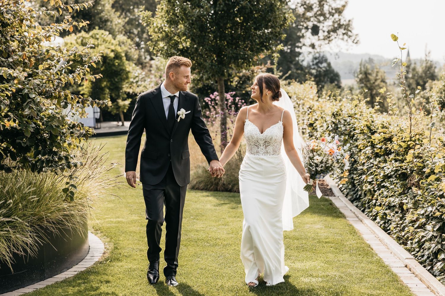 Happy bride and groom walk together through a sunlit garden holding hands on their wedding day.