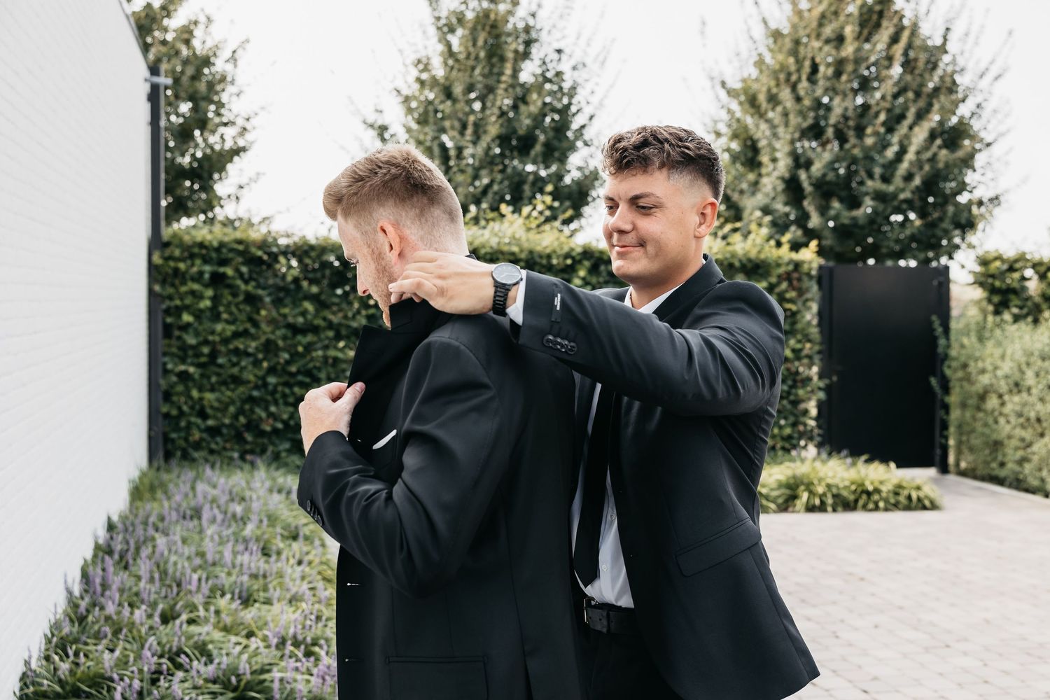 A smiling groomsman adjusts the collar of another mans black suit jacket outdoors.