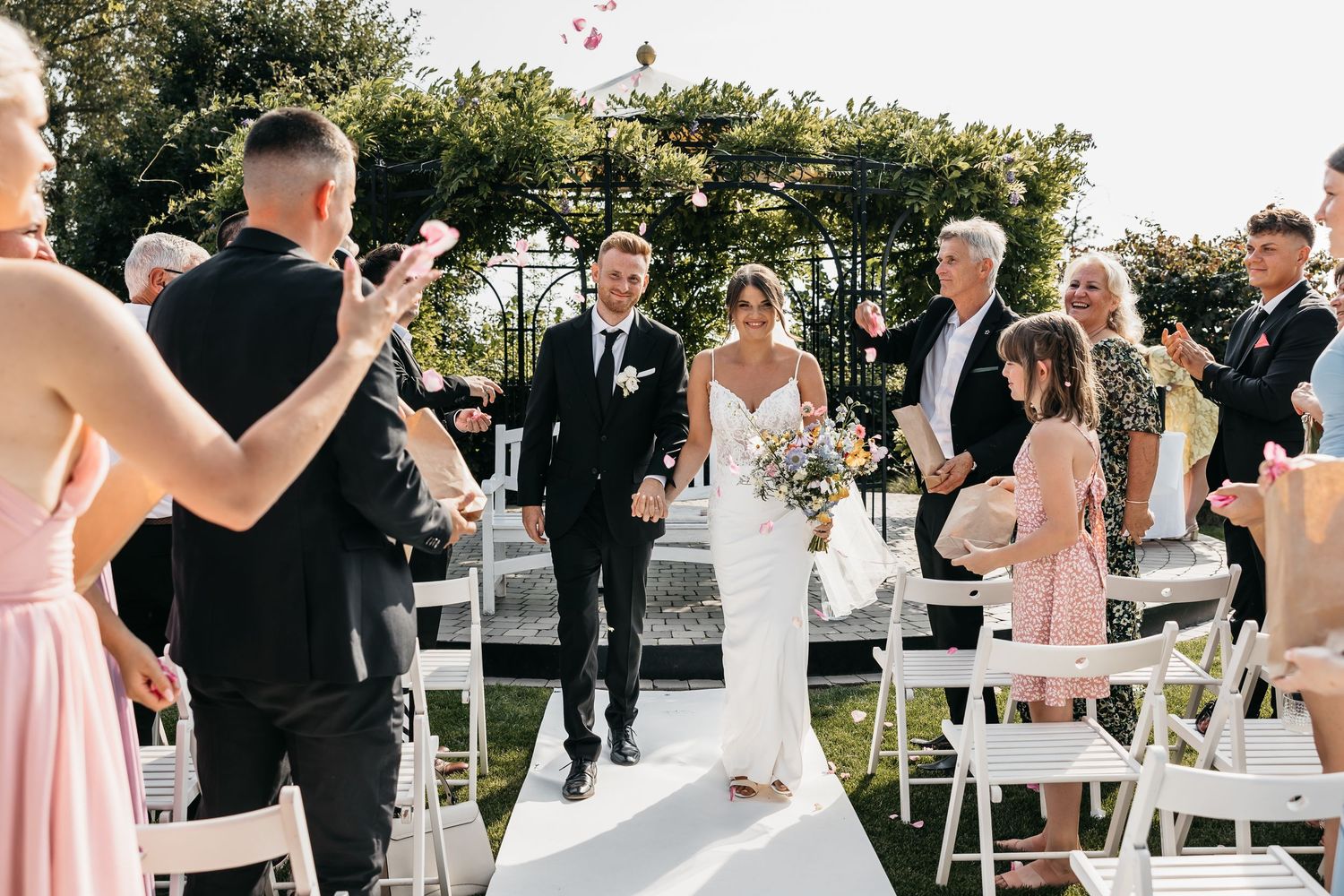 Smiling bride and groom exit outdoor wedding ceremony while guests celebrate with flower confetti.