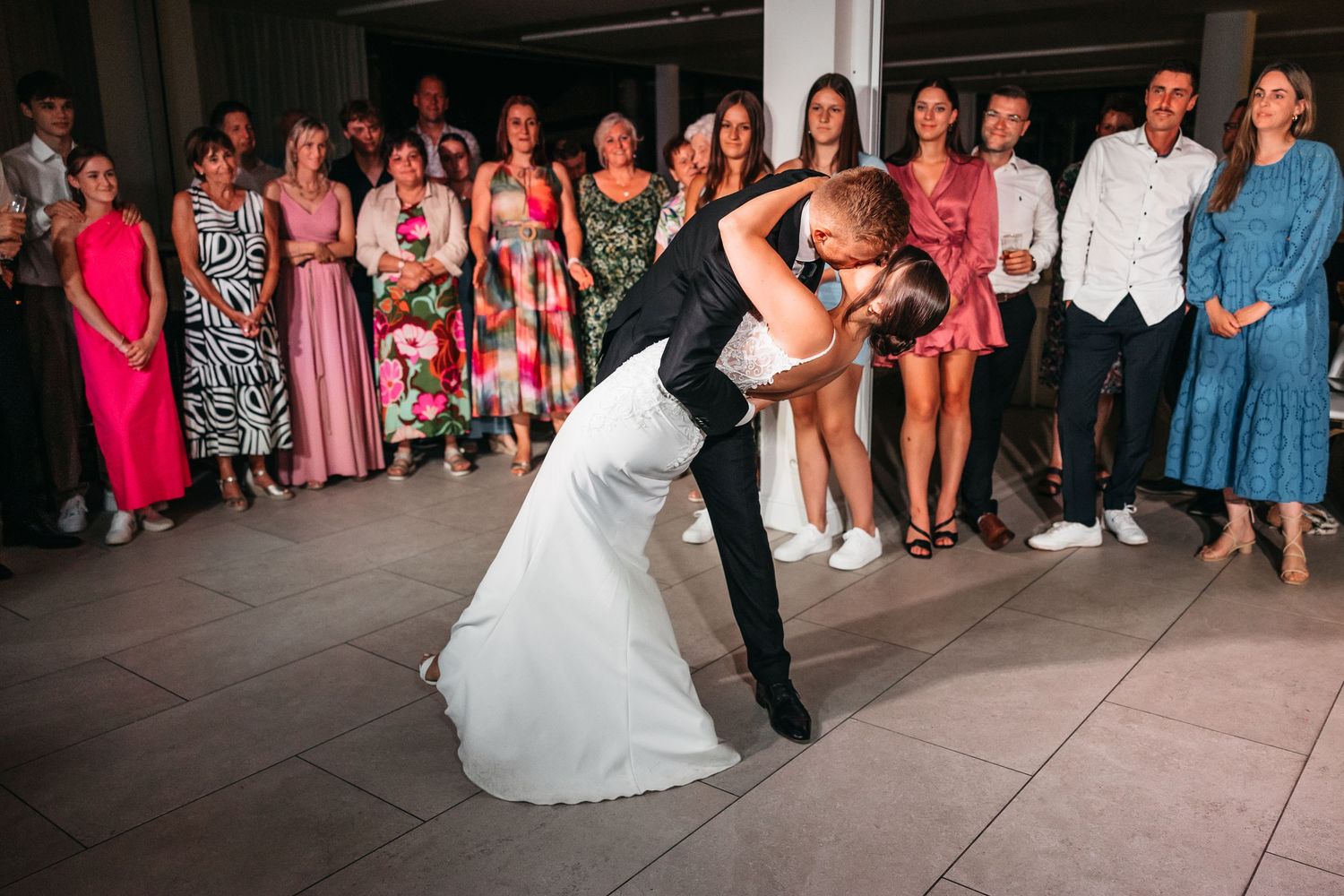 Groom dips bride in stunning white gown during first dance while wedding guests cheer.