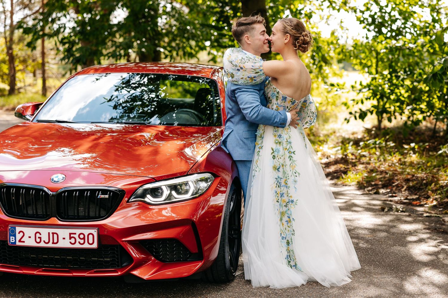 A bride and groom share a tender kiss beside a red BMW M2 sports car surrounded by lush greenery.