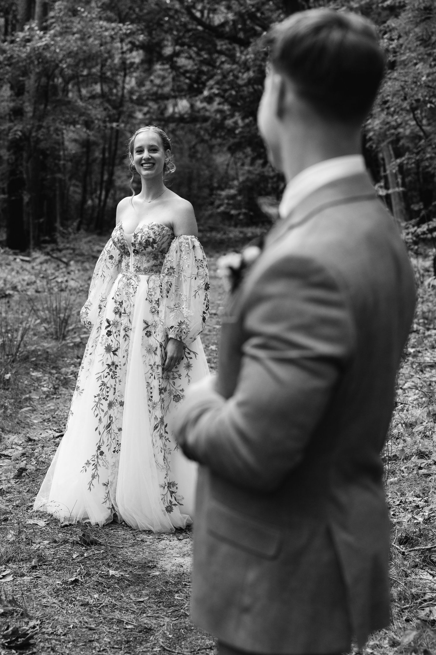Black and white photo of smiling bride in floral gown facing groom on a woodland path.