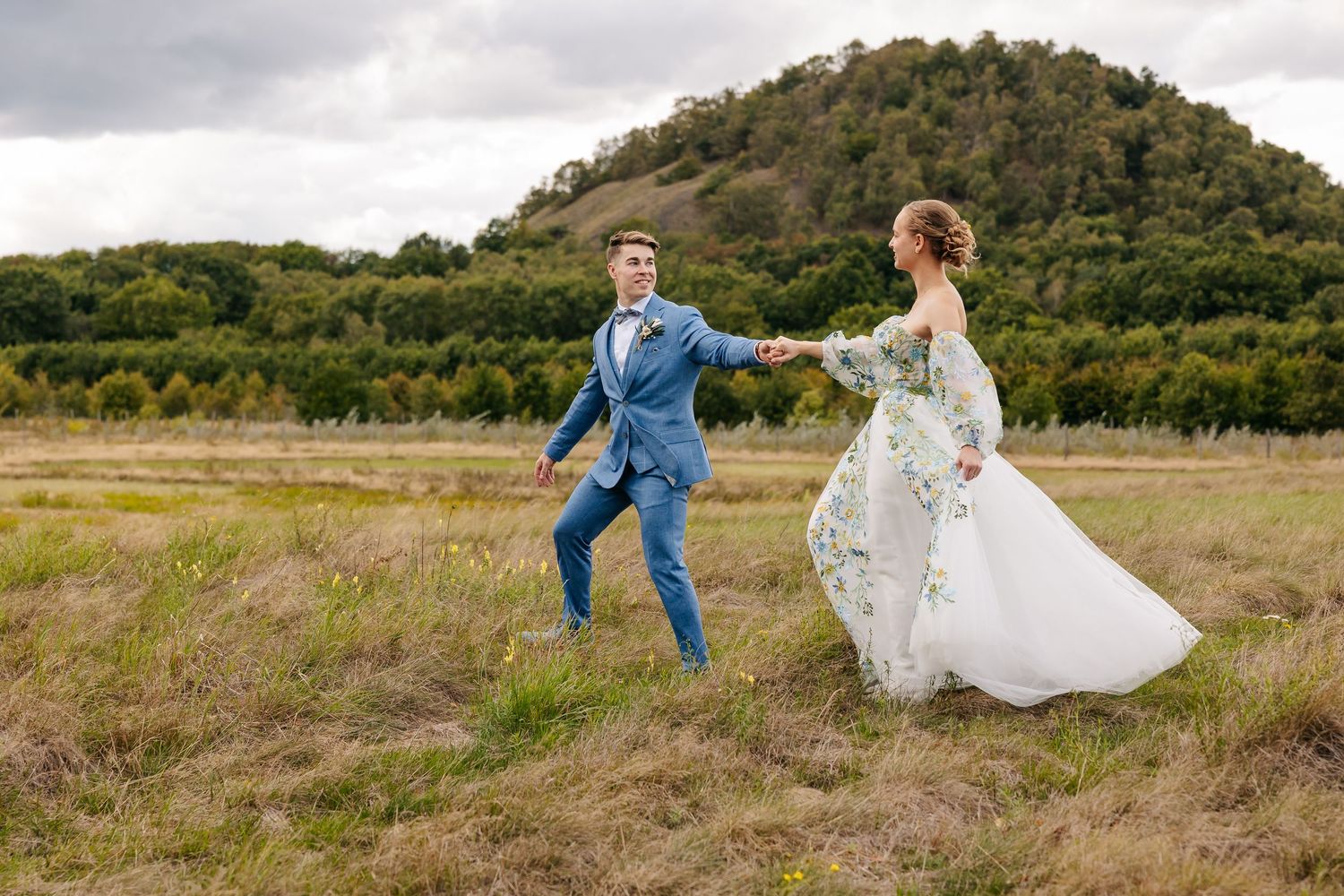 Bride and groom walking hand in hand through grassy field with hill backdrop.
