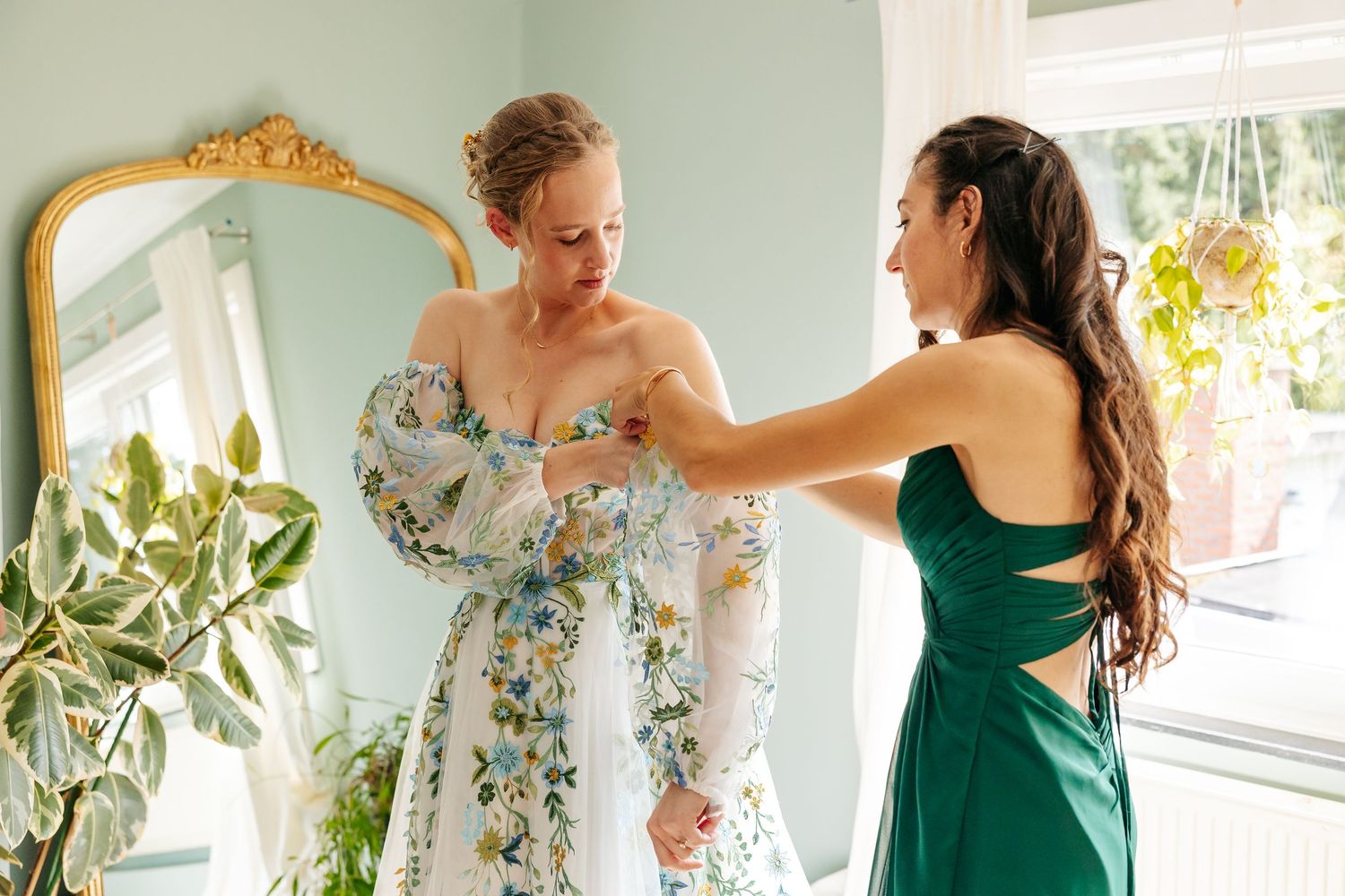 Bridesmaid in green dress helping bride into floral wedding gown before ceremony.