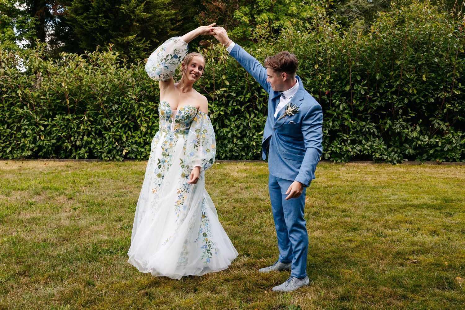 Bride in floral gown twirls with groom in blue suit outdoors on green lawn.