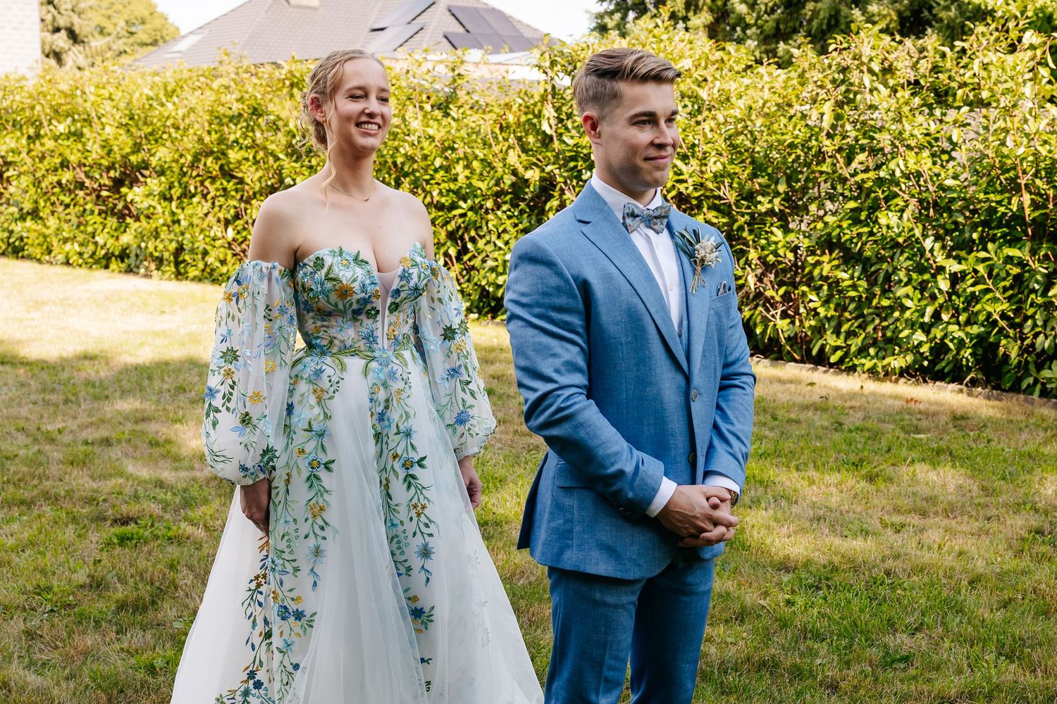 Bride and groom smiling together outdoors, she in floral gown, he in blue suit with bow tie.