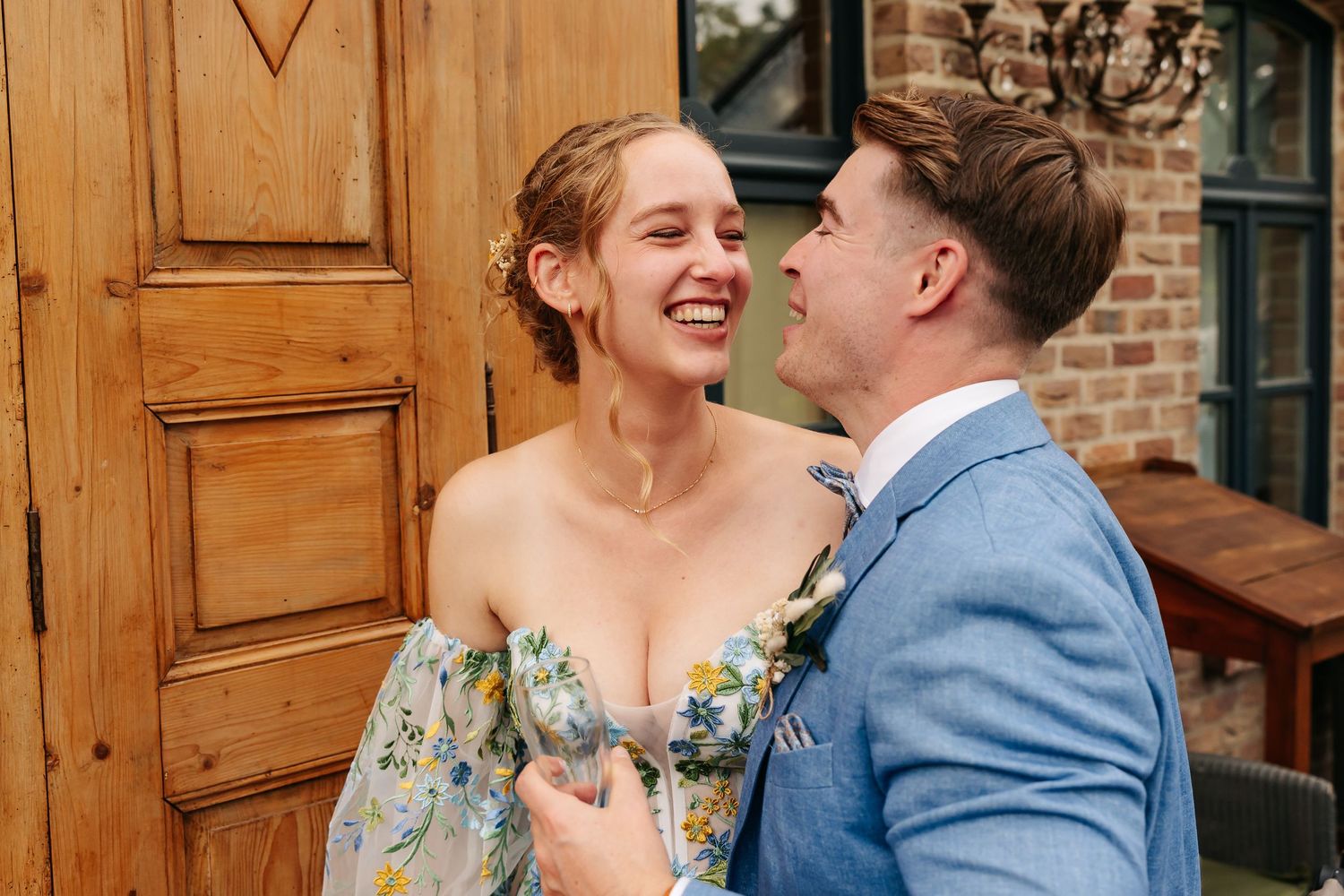 Bride in floral dress and groom in blue suit laughing together outside wooden door at wedding.