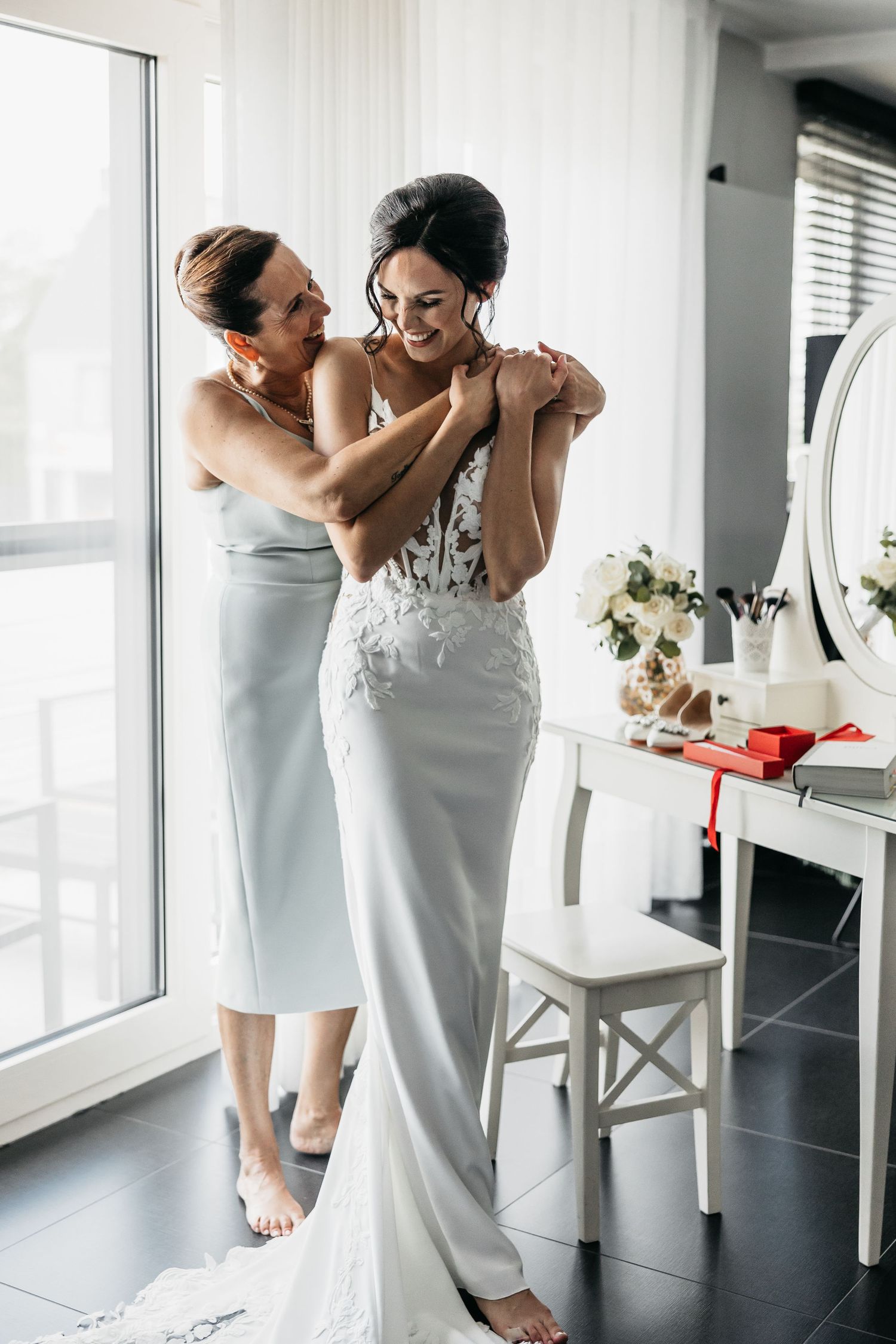Mother helping smiling bride with necklace in a bright wedding preparation room.