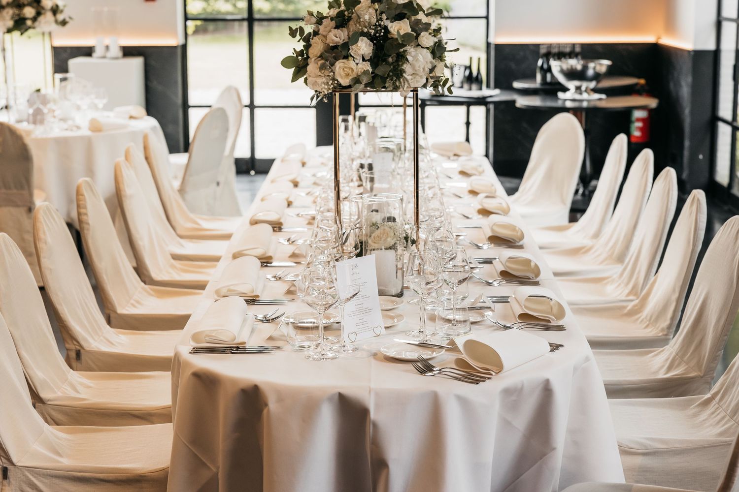 Long wedding reception table with white linens, folded napkins, crystal glasses, and floral centerpieces.