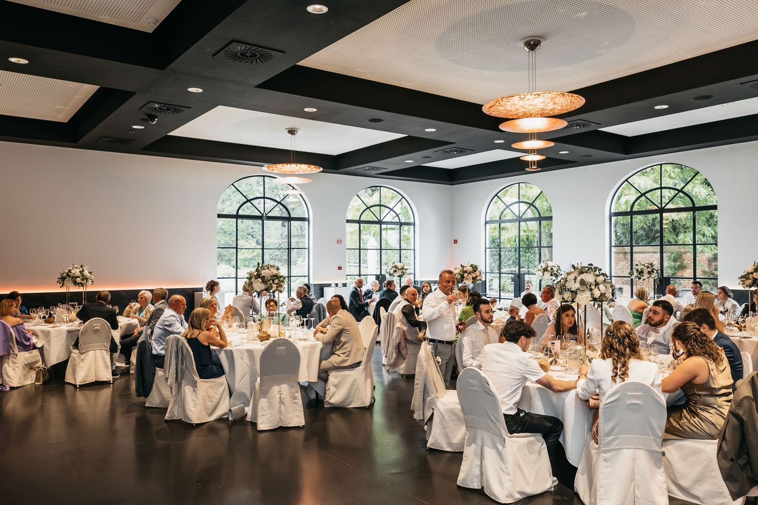 Elegant wedding reception hall with white chair covers, floral centerpieces, and arched windows filling the room with natural light.
