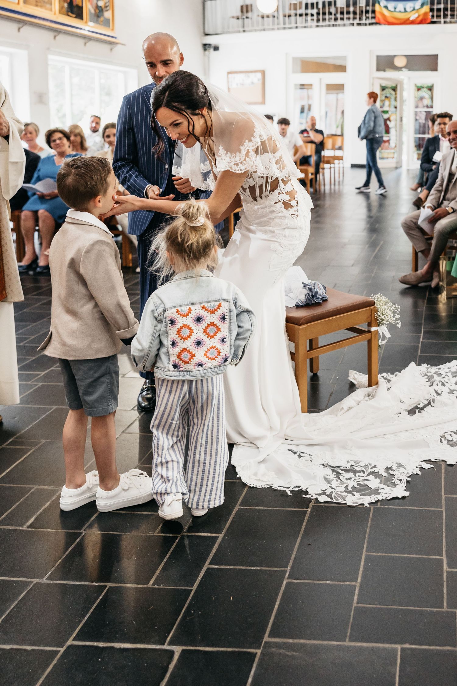 Bride in lace wedding dress bending down to greet two young children during wedding ceremony in church.