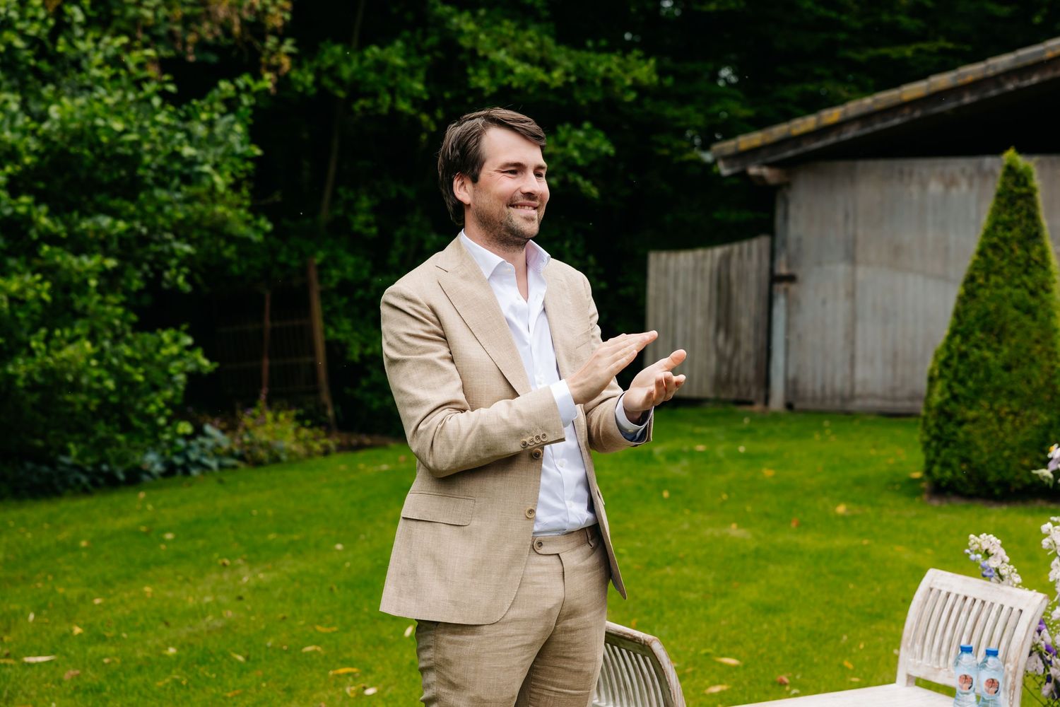 Smiling groom in a beige suit clapping outdoors in a lush green garden setting.