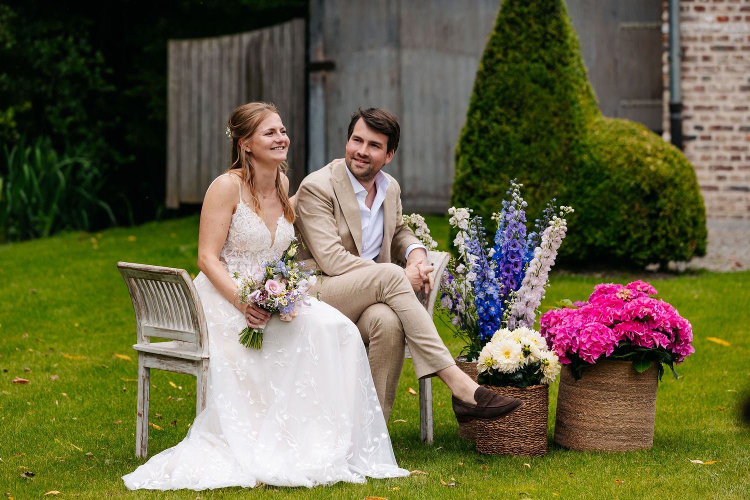 Bride and groom sitting together on a white bench surrounded by colorful flower arrangements in a lush garden.