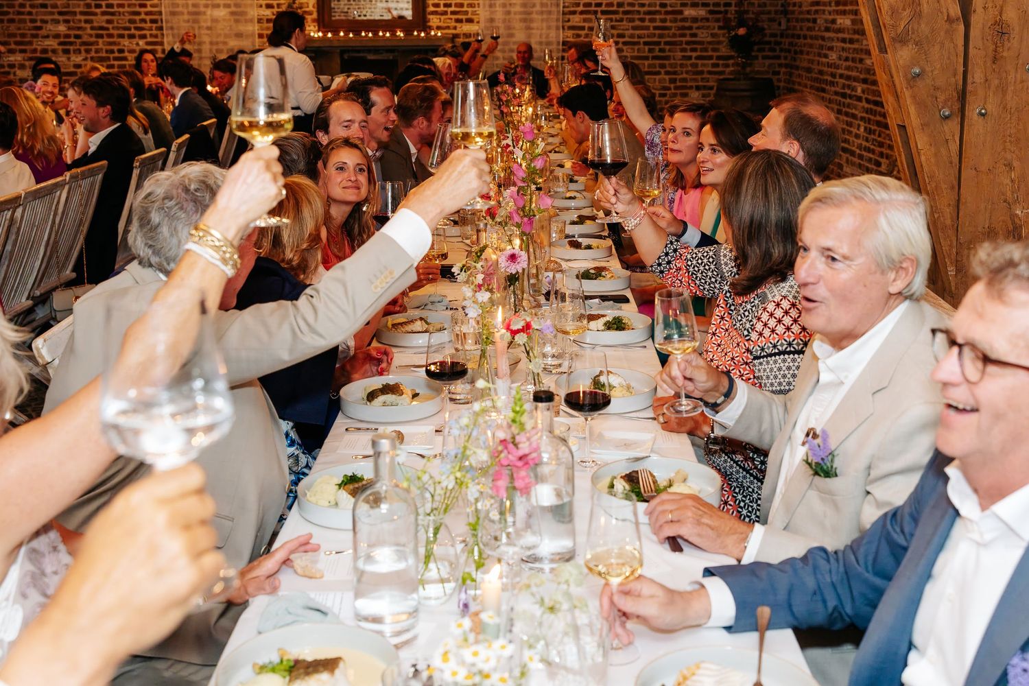 Wedding guests raise champagne glasses in a toast at a long decorated dinner table in a barn venue.