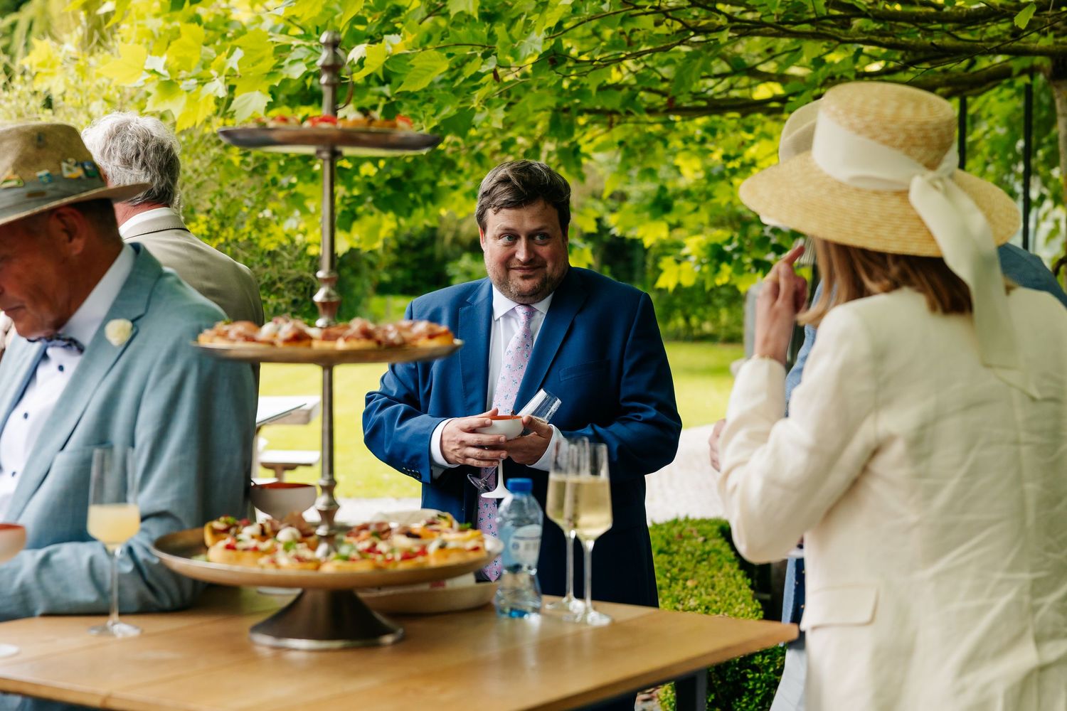 Wedding guests enjoying champagne and appetizers at an outdoor reception under lush green trees.