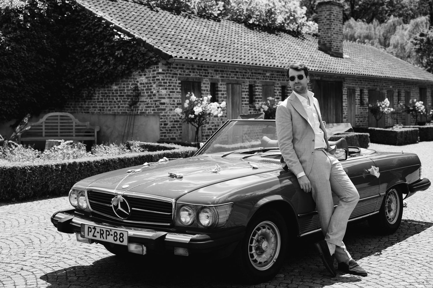 Vintage black and white photo of man in white suit leaning on classic Mercedes convertible.