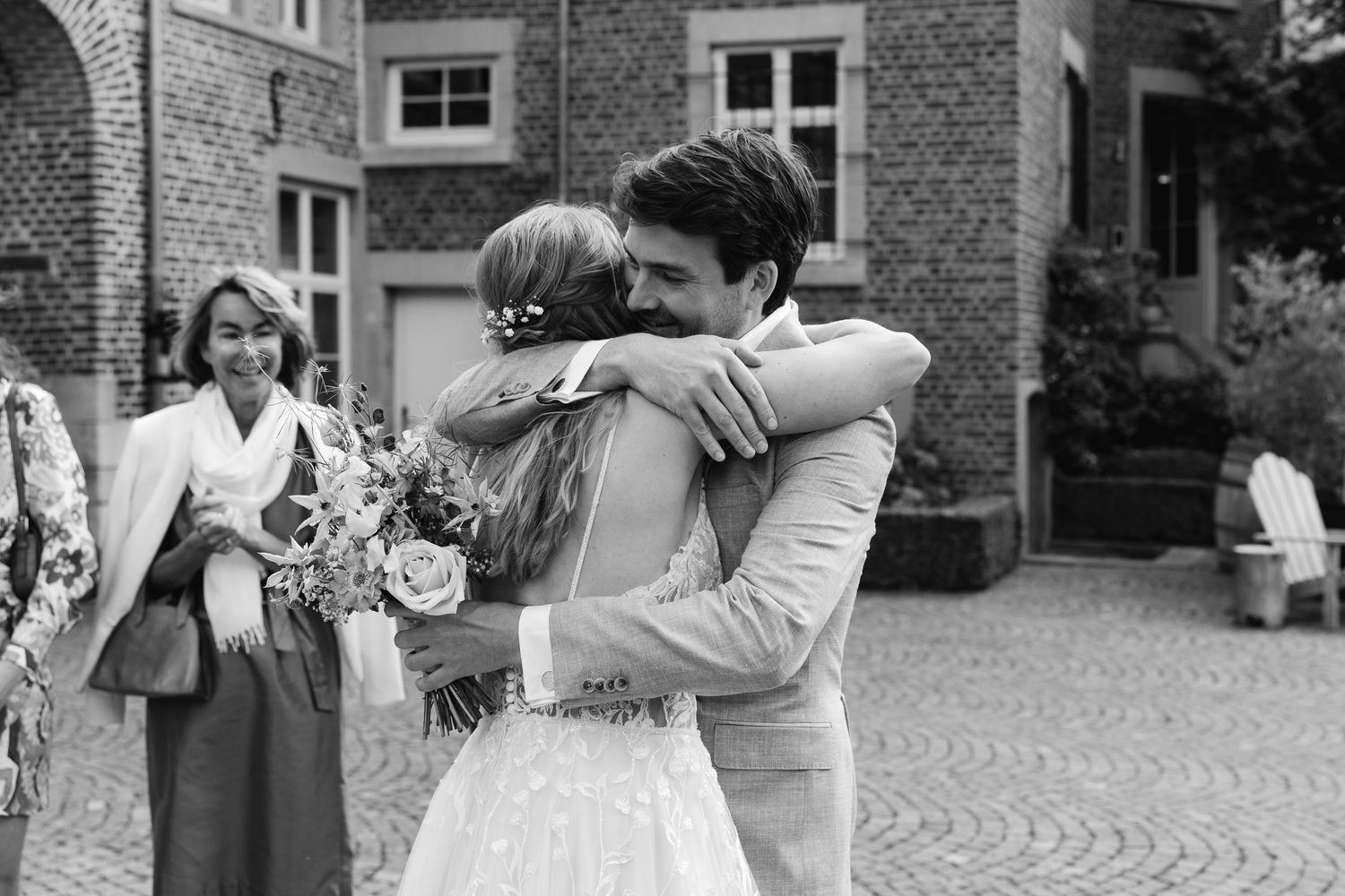 Black and white photo of groom hugging bride in lace dress with guests smiling in background.