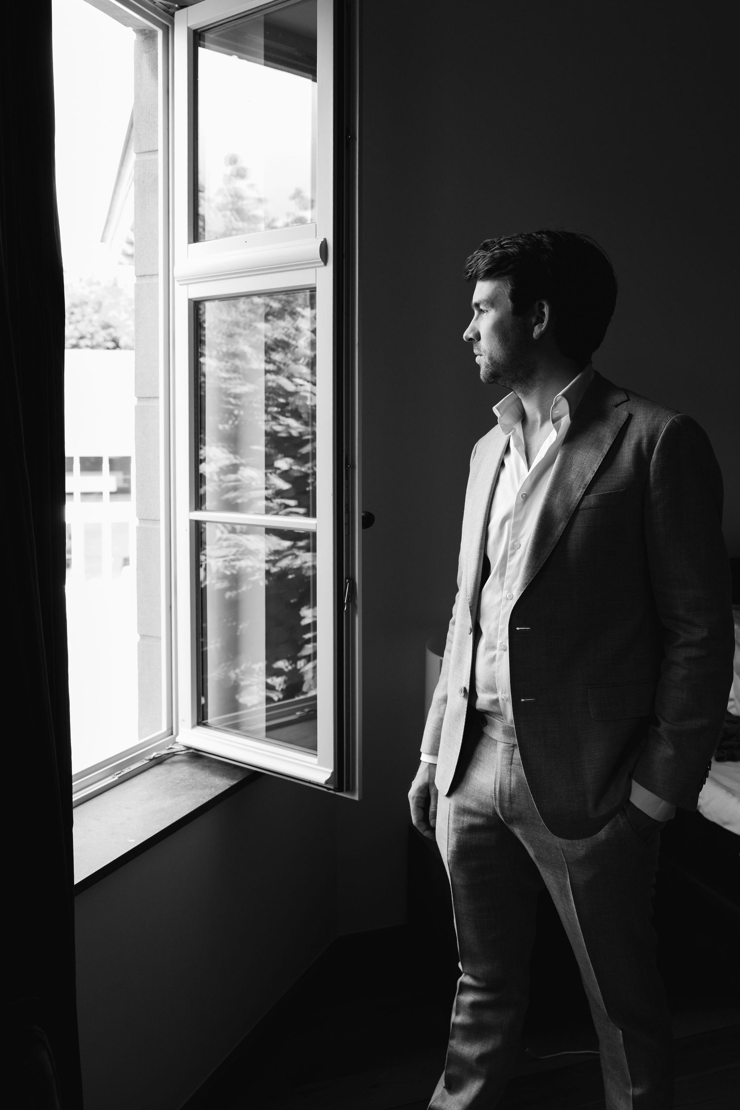 Black and white photo of a stylish man in a suit gazing out an open window with natural light.