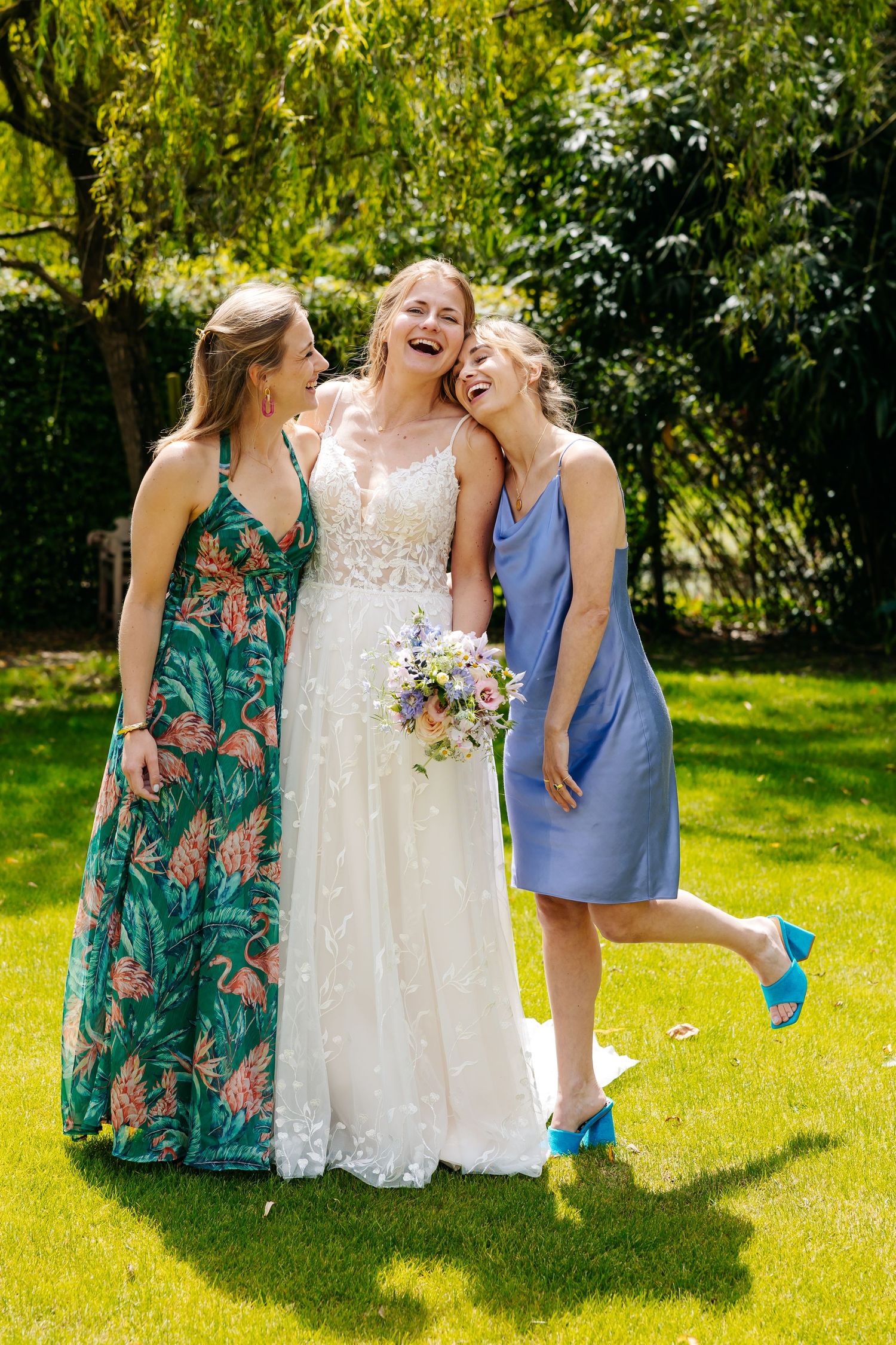 Bride in white gown laughing with two friends in colorful dresses on sunny green lawn.