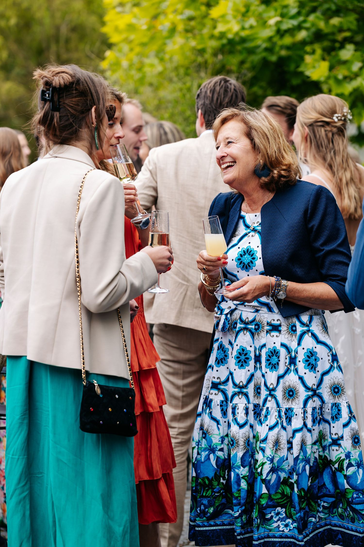 Smiling wedding guests in colorful dresses toast with champagne glasses at outdoor garden reception.