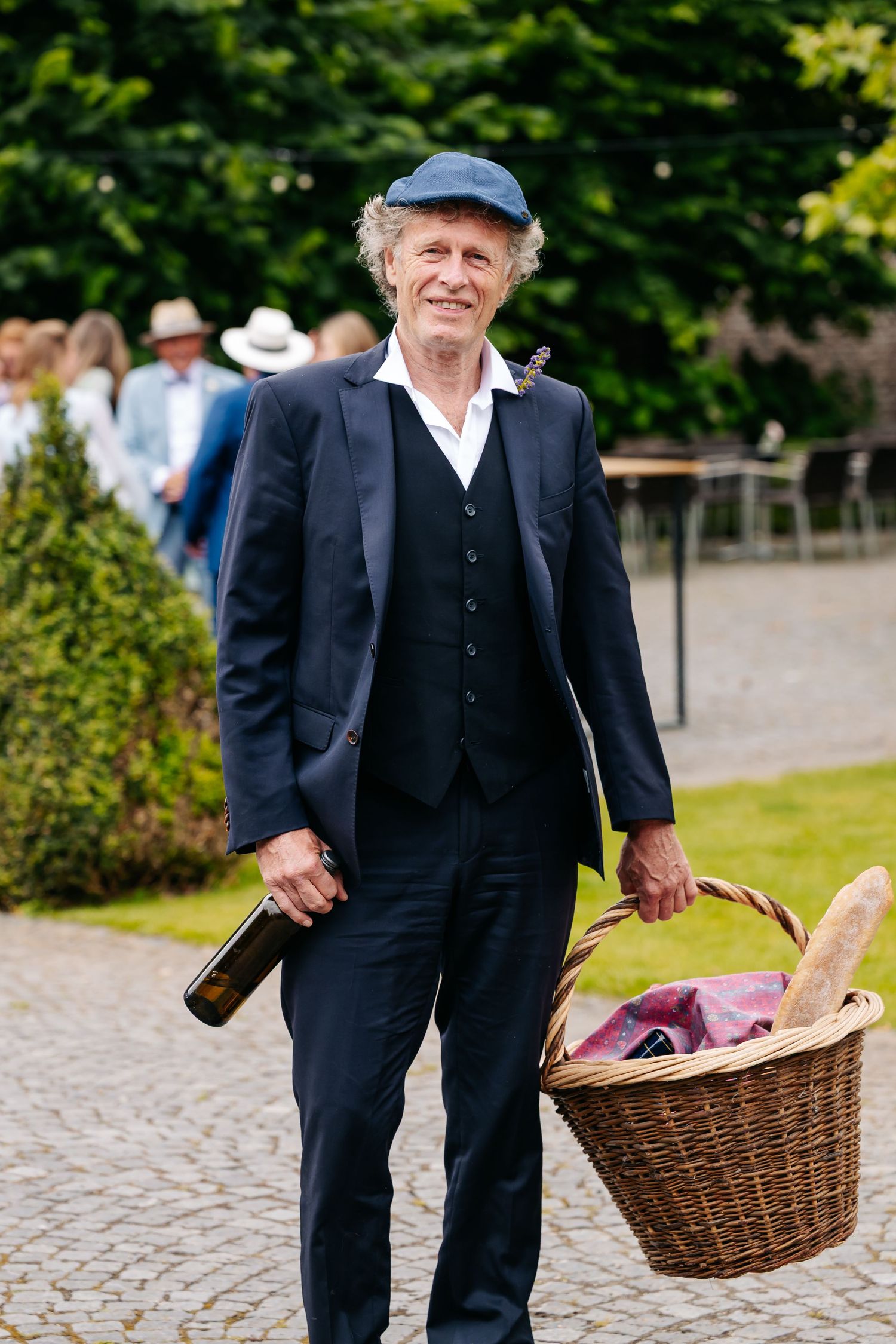 Elderly man in navy suit and flat cap carries wicker picnic basket and wine bottle at outdoor event.