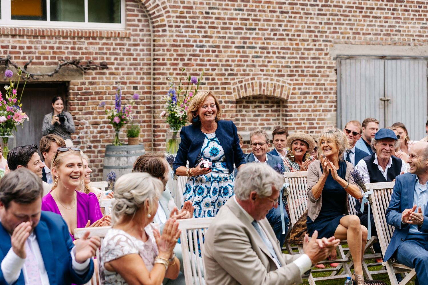 Wedding guests laugh and celebrate at outdoor reception against rustic brick wall venue on sunny day.
