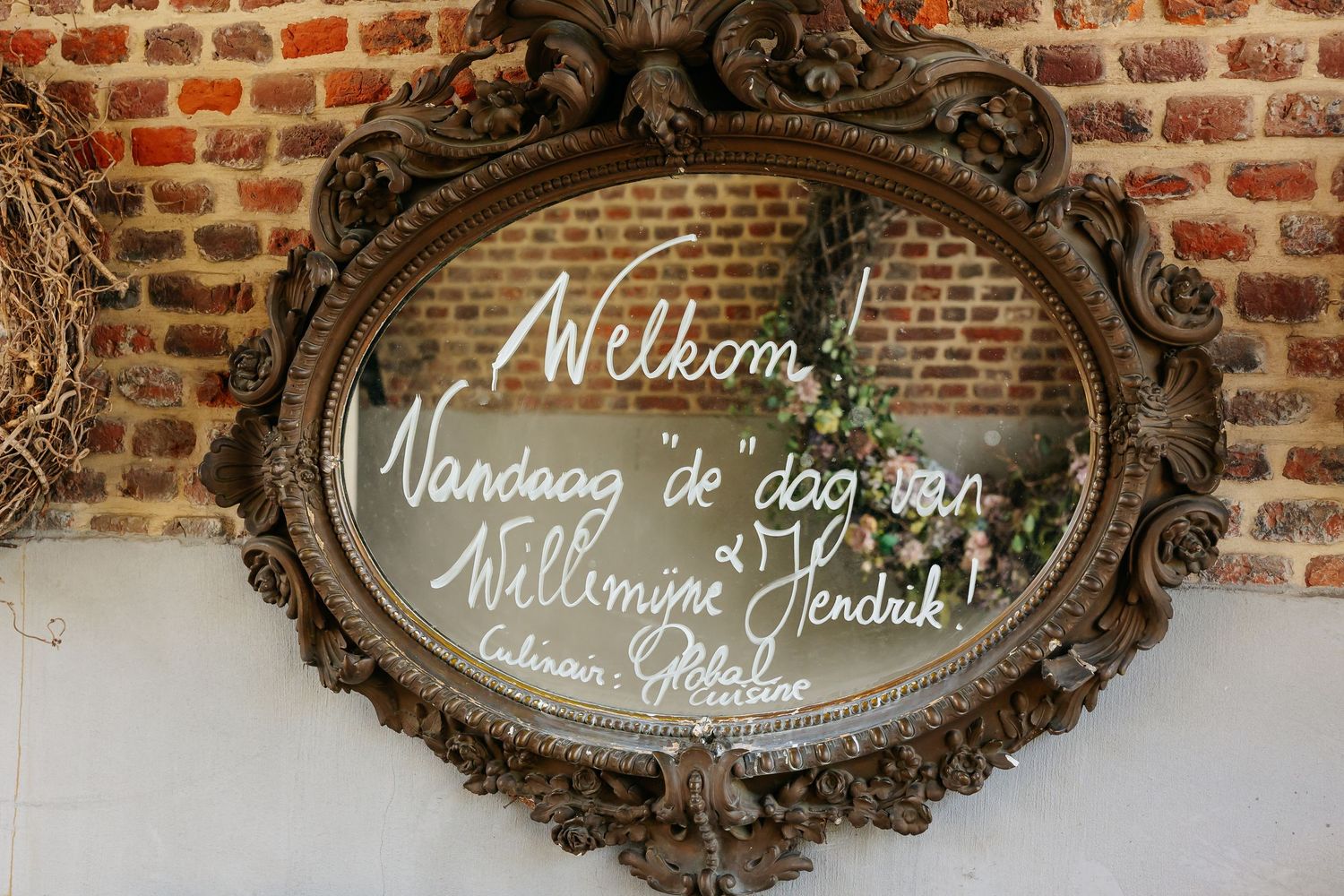 Ornate vintage mirror with Dutch wedding welcome message displayed against a rustic brick wall with floral decor.