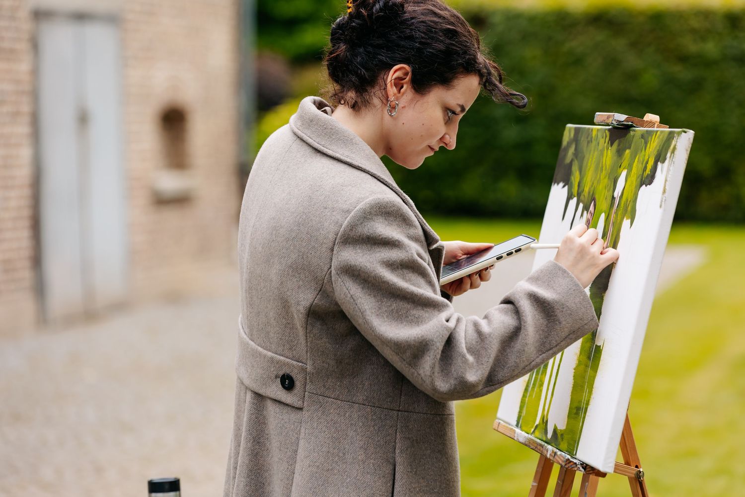 Artist painting on an easel outdoors beside a rustic brick building in a garden setting.