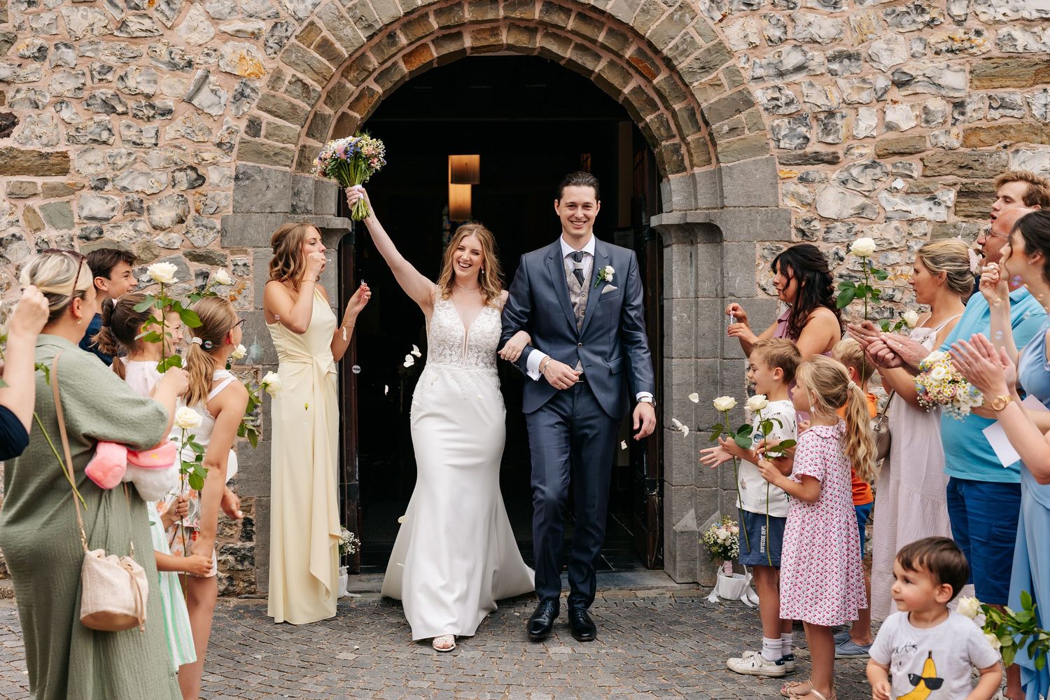 Bride and groom exit stone church to cheering guests throwing confetti on their wedding day.