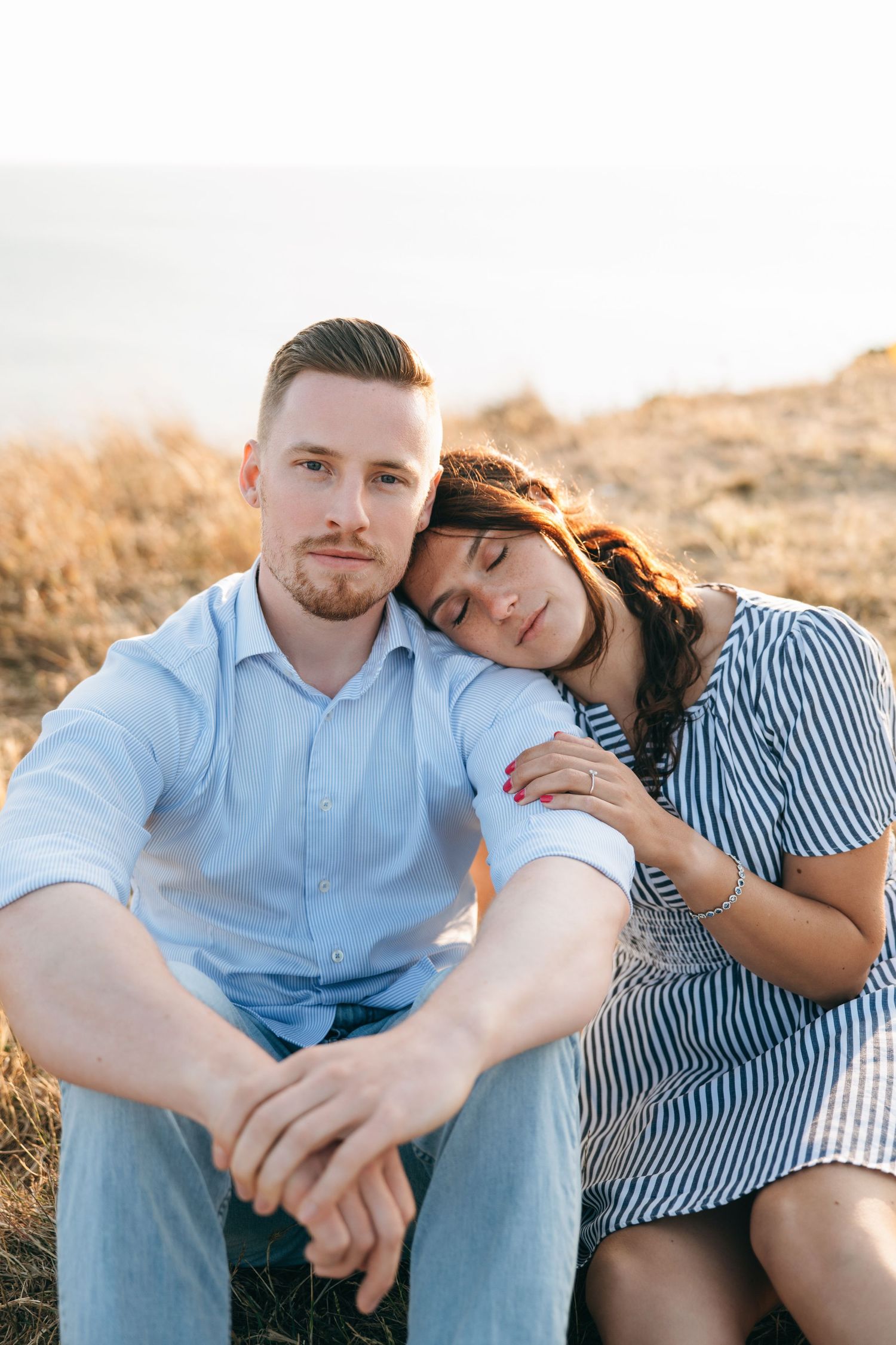 Couple sitting outdoors at sunset, woman resting head on mans shoulder in tender romantic moment.