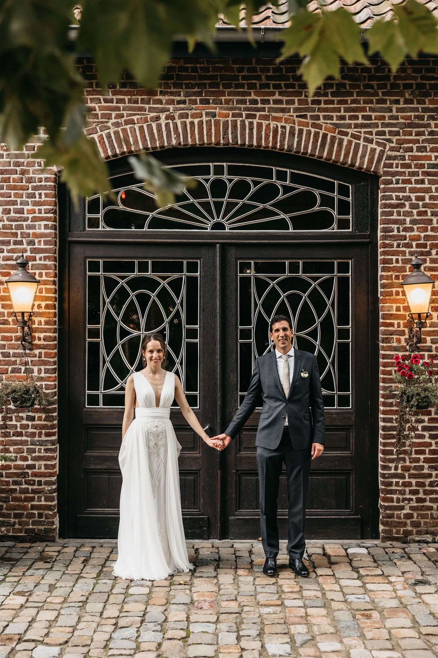Bride and groom holding hands before ornate dark wooden doors on cobblestone courtyard.
