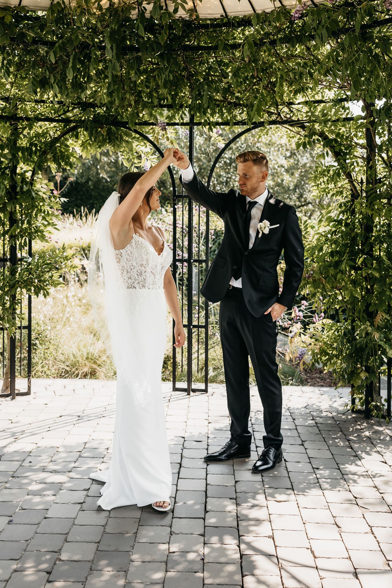 Groom spinning bride under green garden arbor in elegant wedding attire.