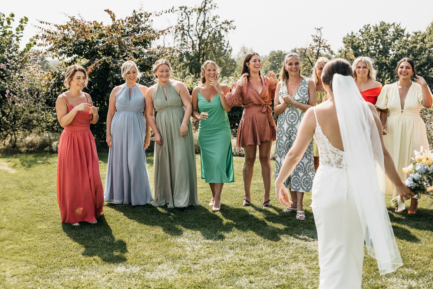 Bride with veil prepares to toss bouquet to group of women in colorful mismatched dresses at outdoor wedding.