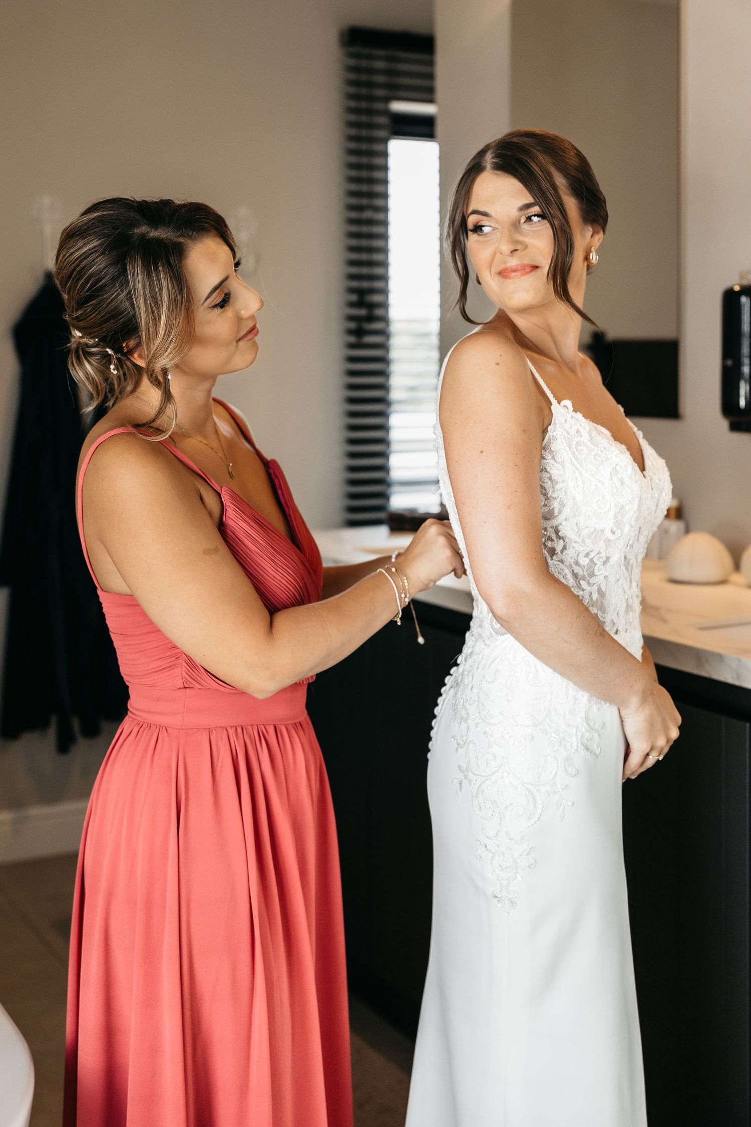 Bridesmaid in coral dress zipping up bride's white lace wedding gown before ceremony.