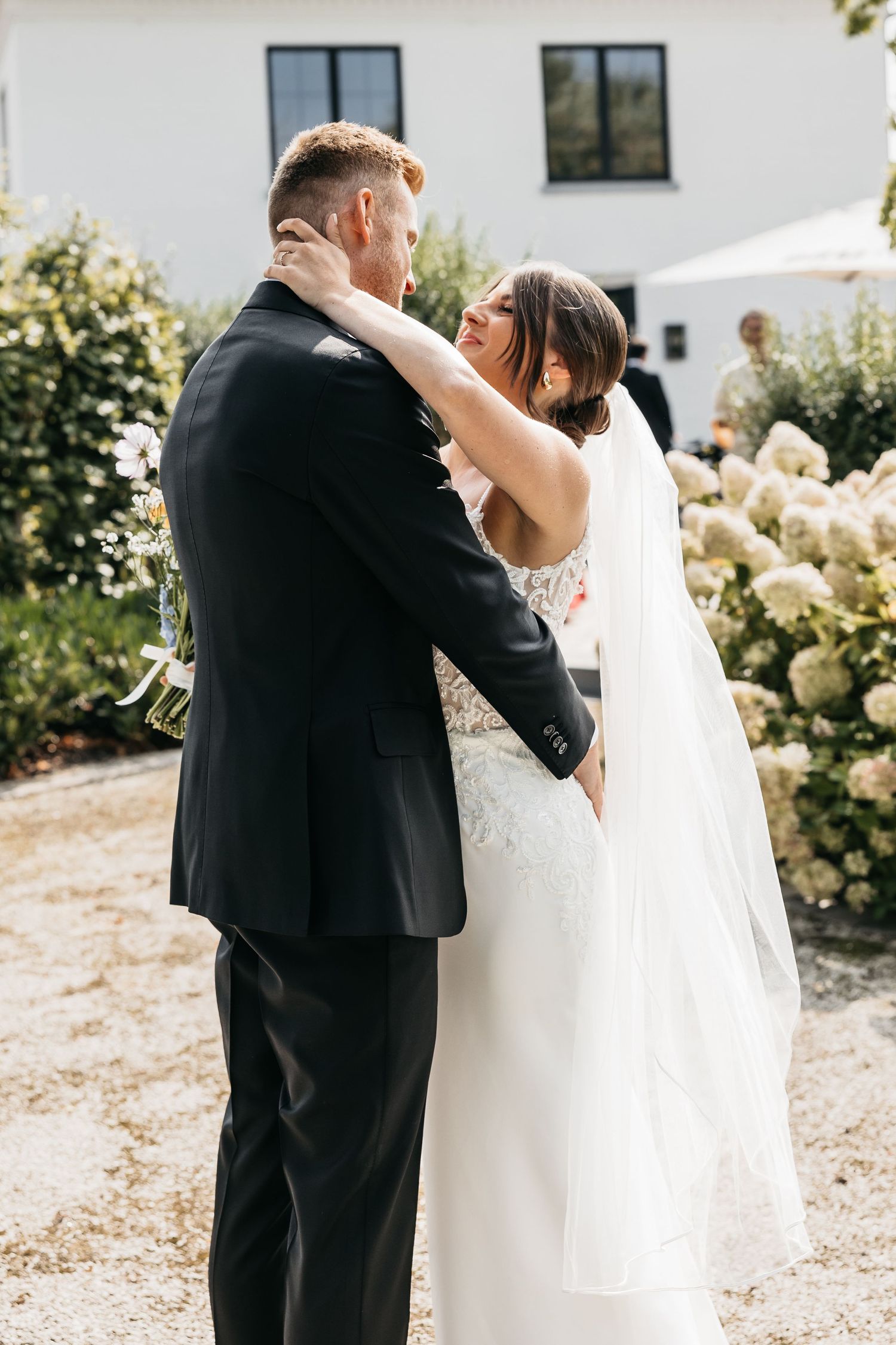 Bride and groom share tender embrace during outdoor first look at elegant white venue.