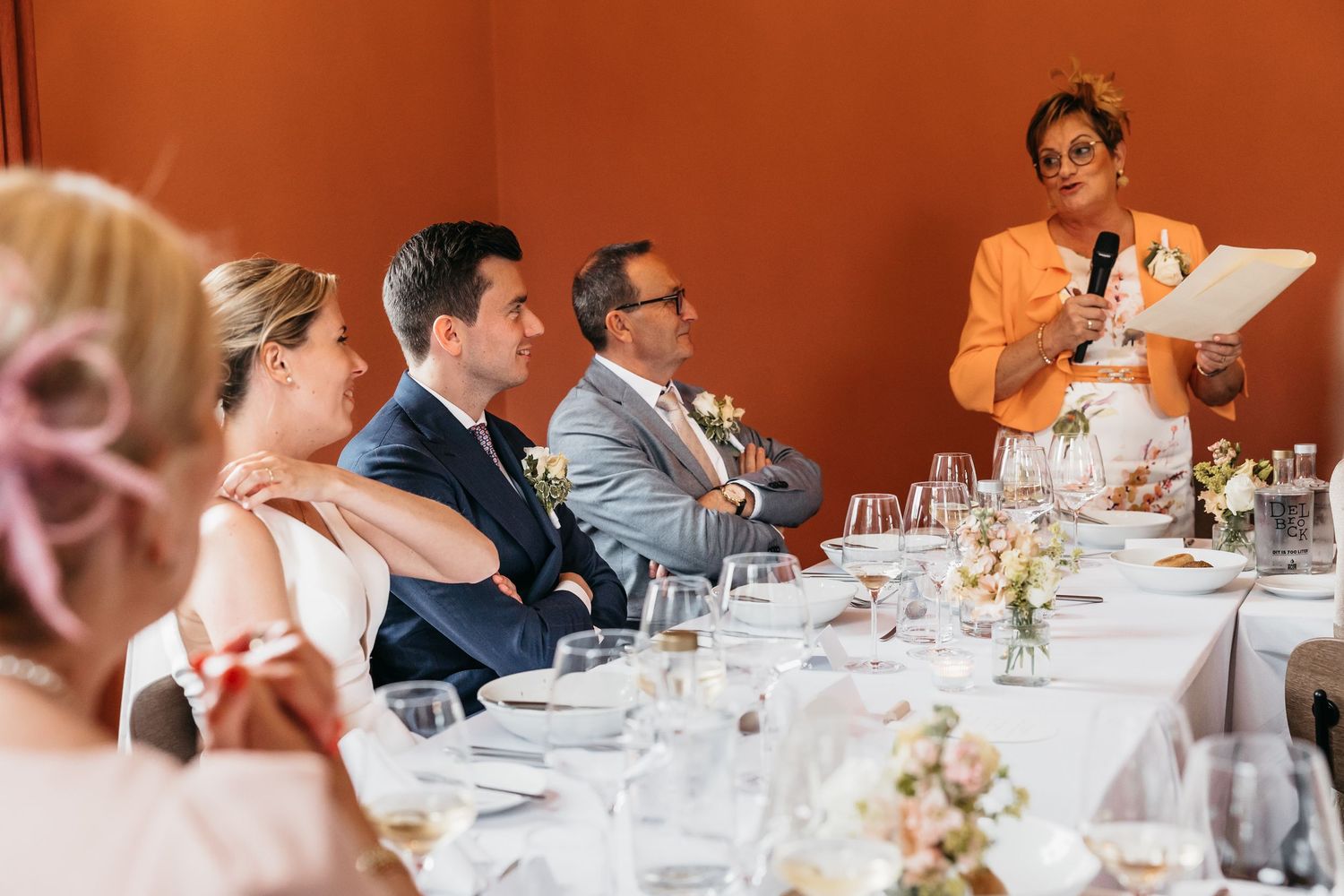 Guest reads speech at wedding reception while bride and groom listen at decorated table.