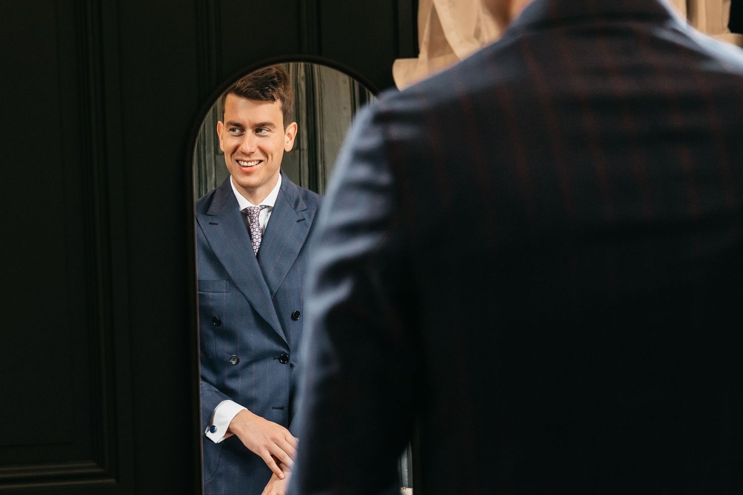 Smiling groom in navy suit adjusting his appearance in an oval mirror.