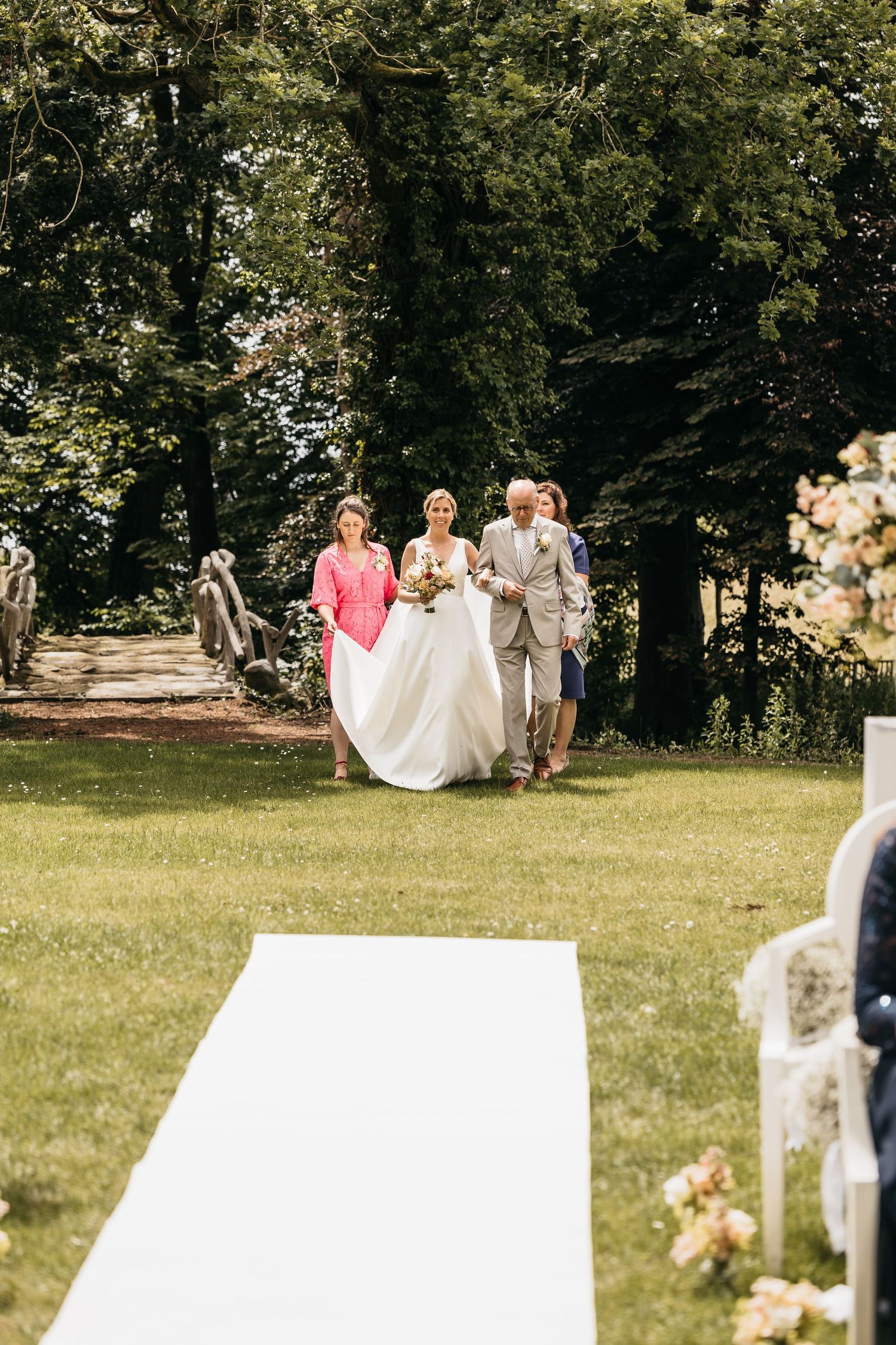 Bride in white gown walks down garden aisle accompanied by father and bridesmaids at outdoor ceremony.