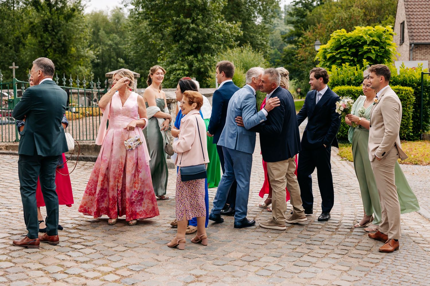 Wedding guests in colorful formal attire greet each other on cobblestone plaza.