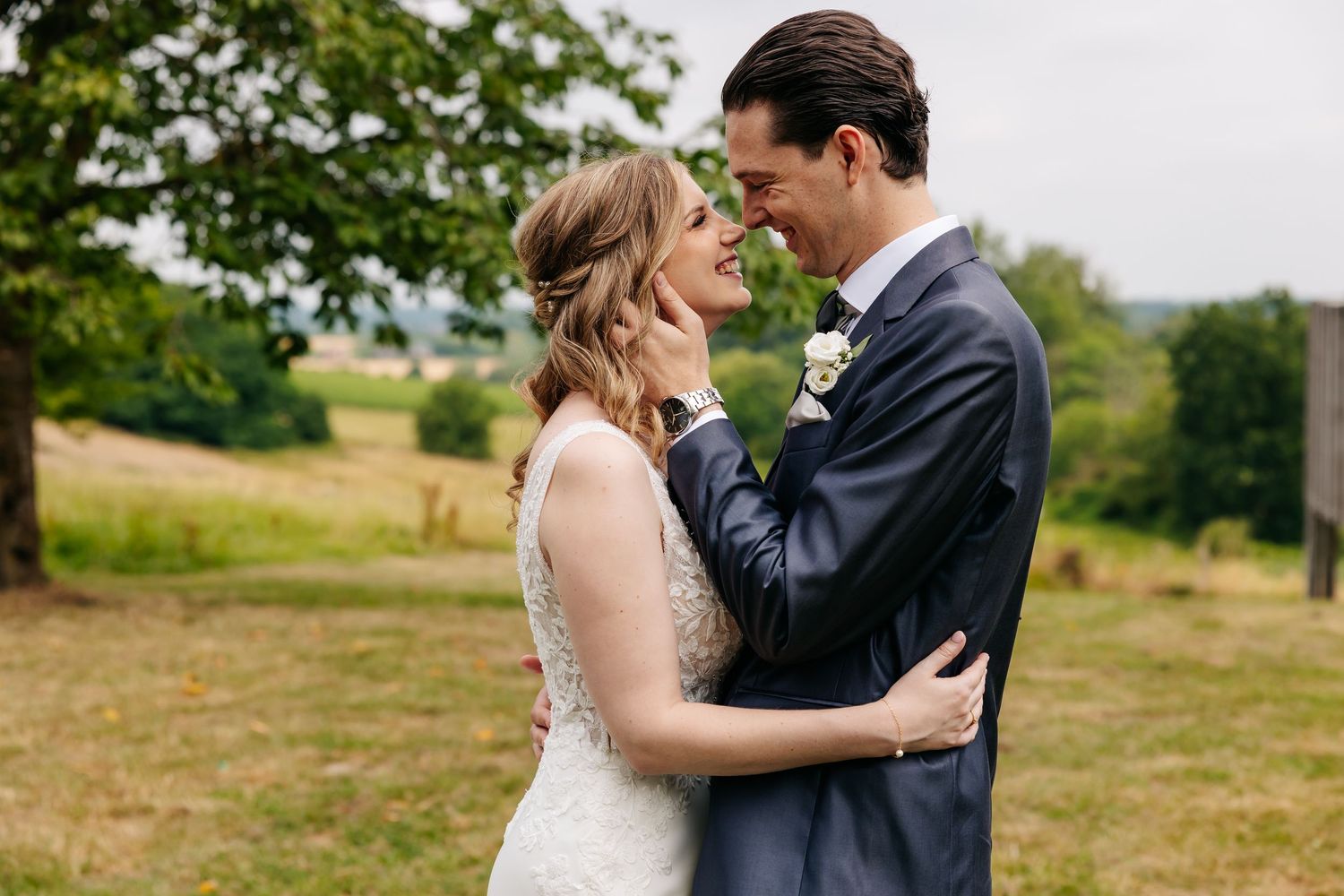 Groom tenderly holding the bride's face as they smile at each other in a sunlit green field.