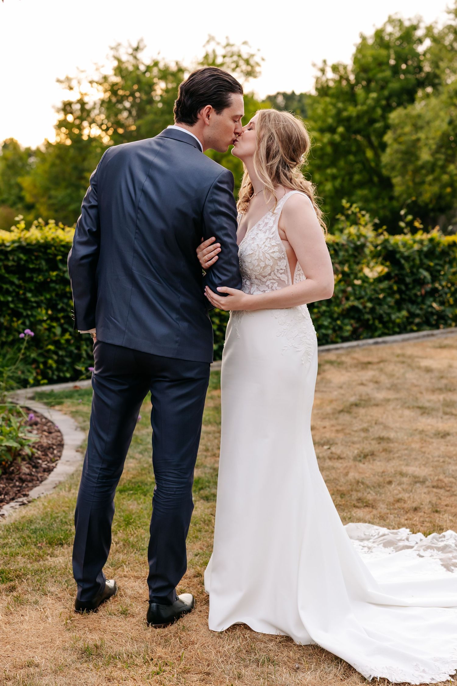 Bride and groom share a kiss in a lush garden, bride wearing a white lace gown with a long train.