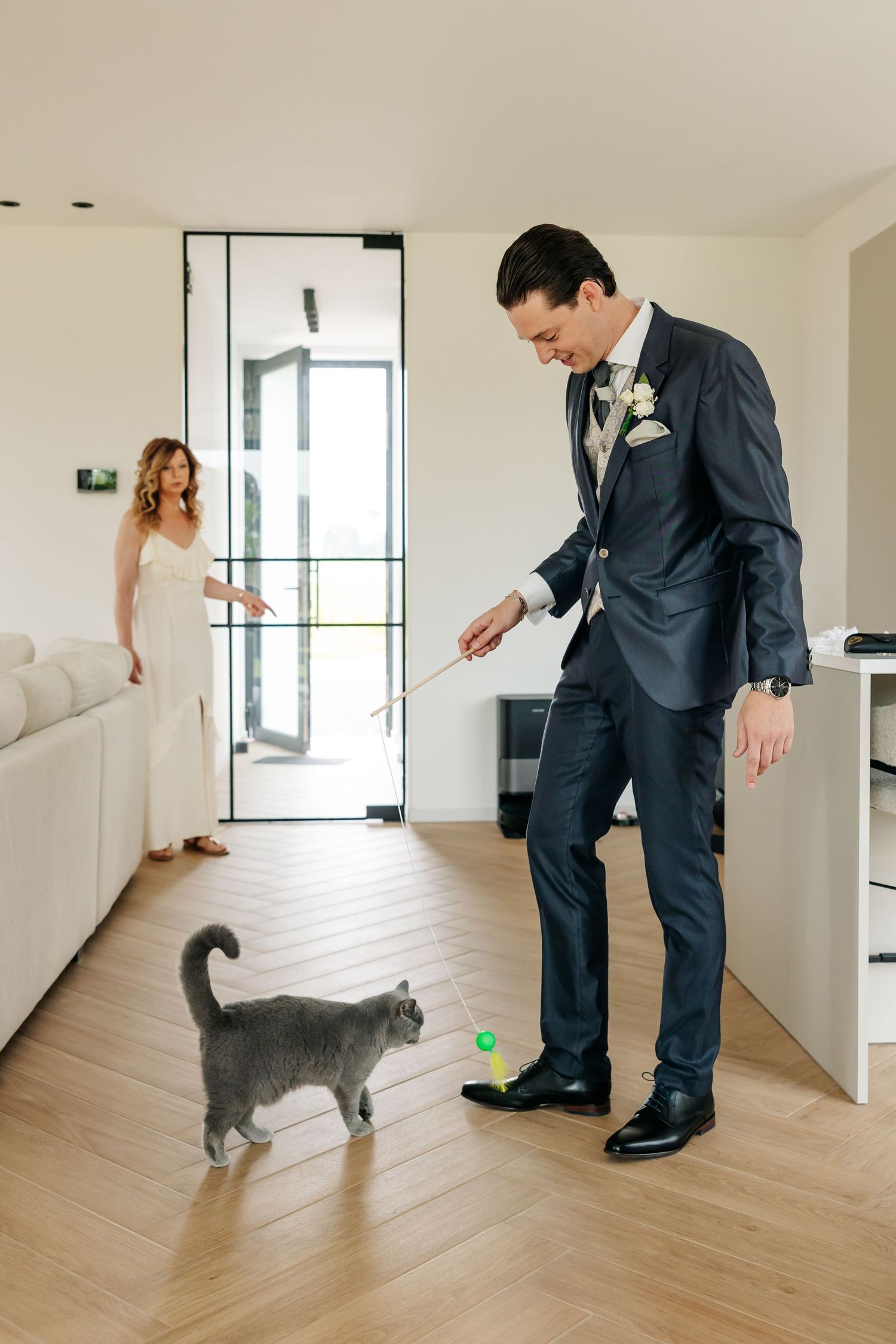 A groom in navy suit plays with gray cat using toy wand while bride watches in bright modern home.