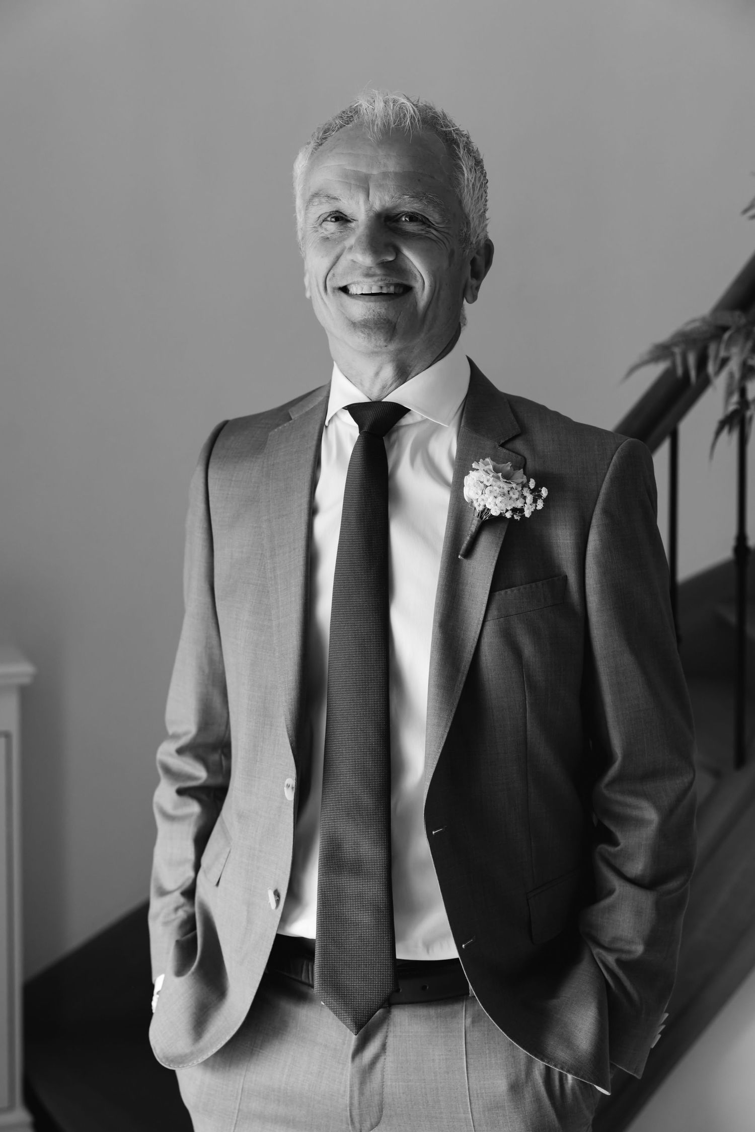Elegant groom in suit with boutonniere smiles confidently near a staircase in black and white photo.