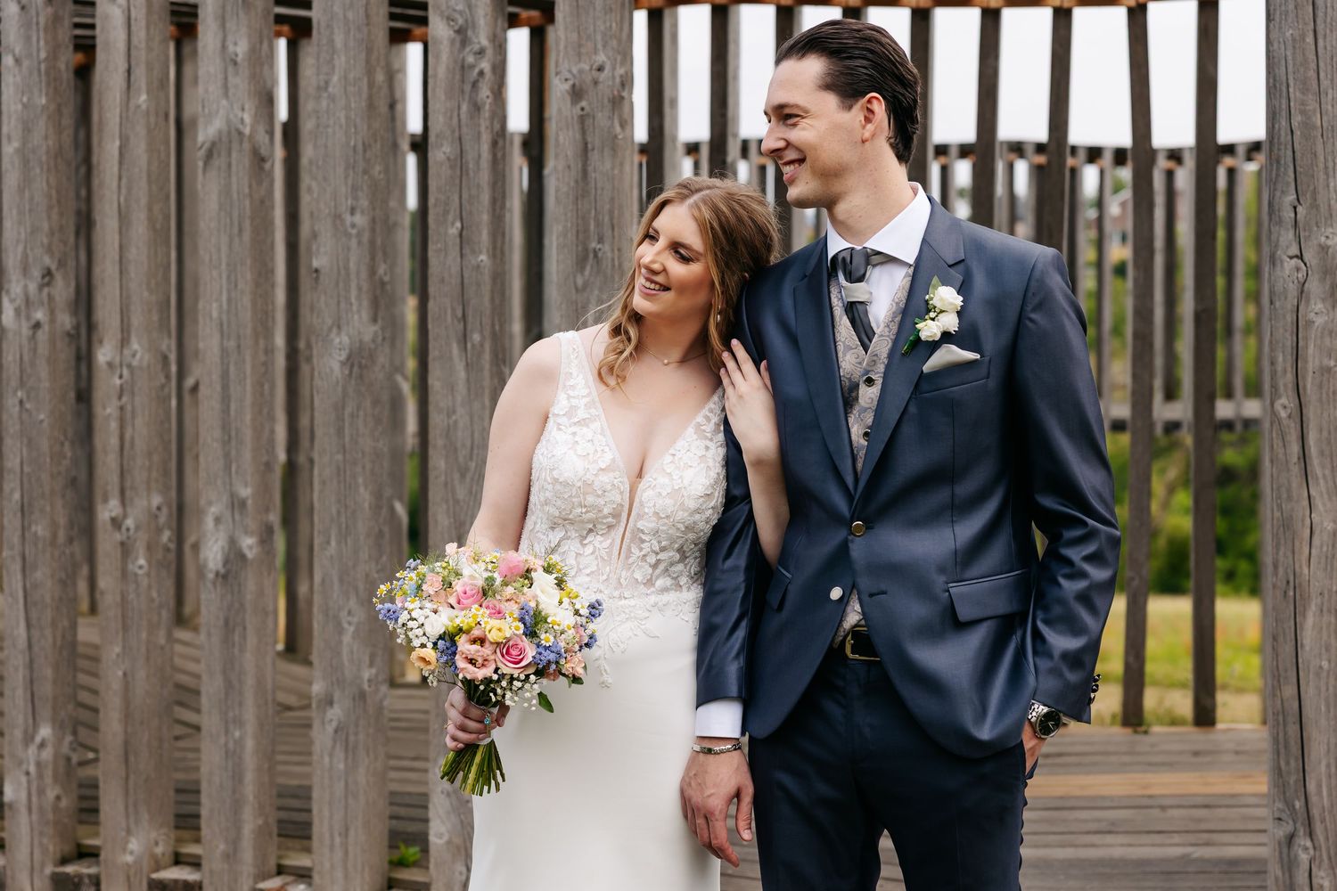 Smiling bride holds a colorful bouquet beside her tall groom in a navy suit near wooden structures.