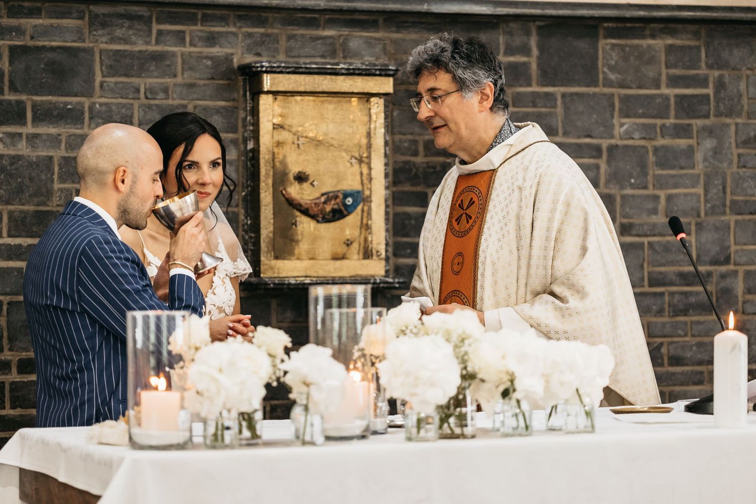 Priest smiling at bride and groom during Catholic wedding ceremony at flower-adorned altar.