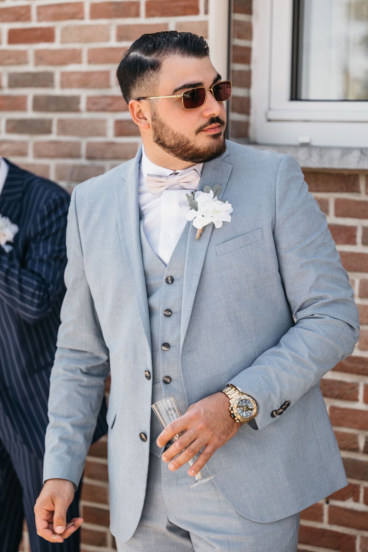 Elegant groom in grey three-piece suit with white boutonniere, bow tie, and luxury watch at wedding.