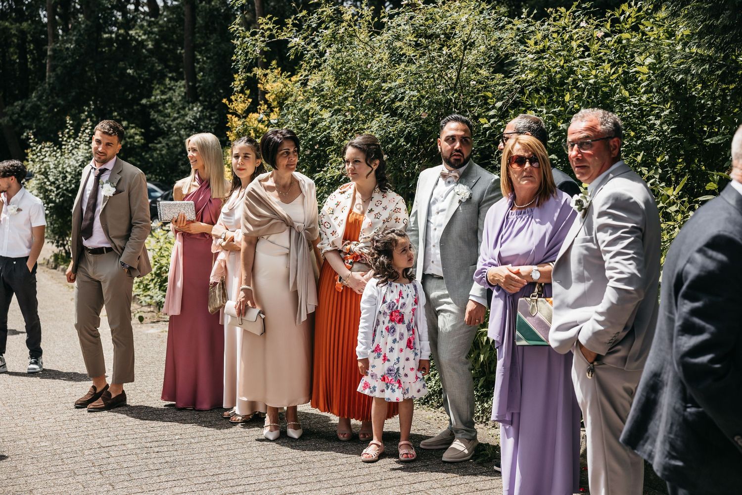 Wedding guests in colorful formal attire standing outdoors in a garden setting on a sunny day.