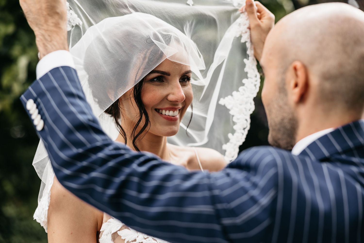 Groom lifts bride's lace veil revealing her radiant smile in a tender wedding moment.