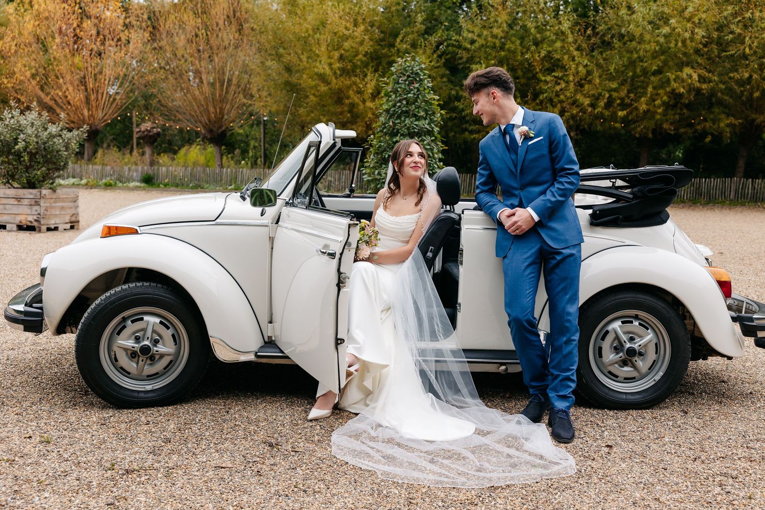 Bride and groom posing beside a classic white VW Beetle convertible surrounded by autumn foliage.